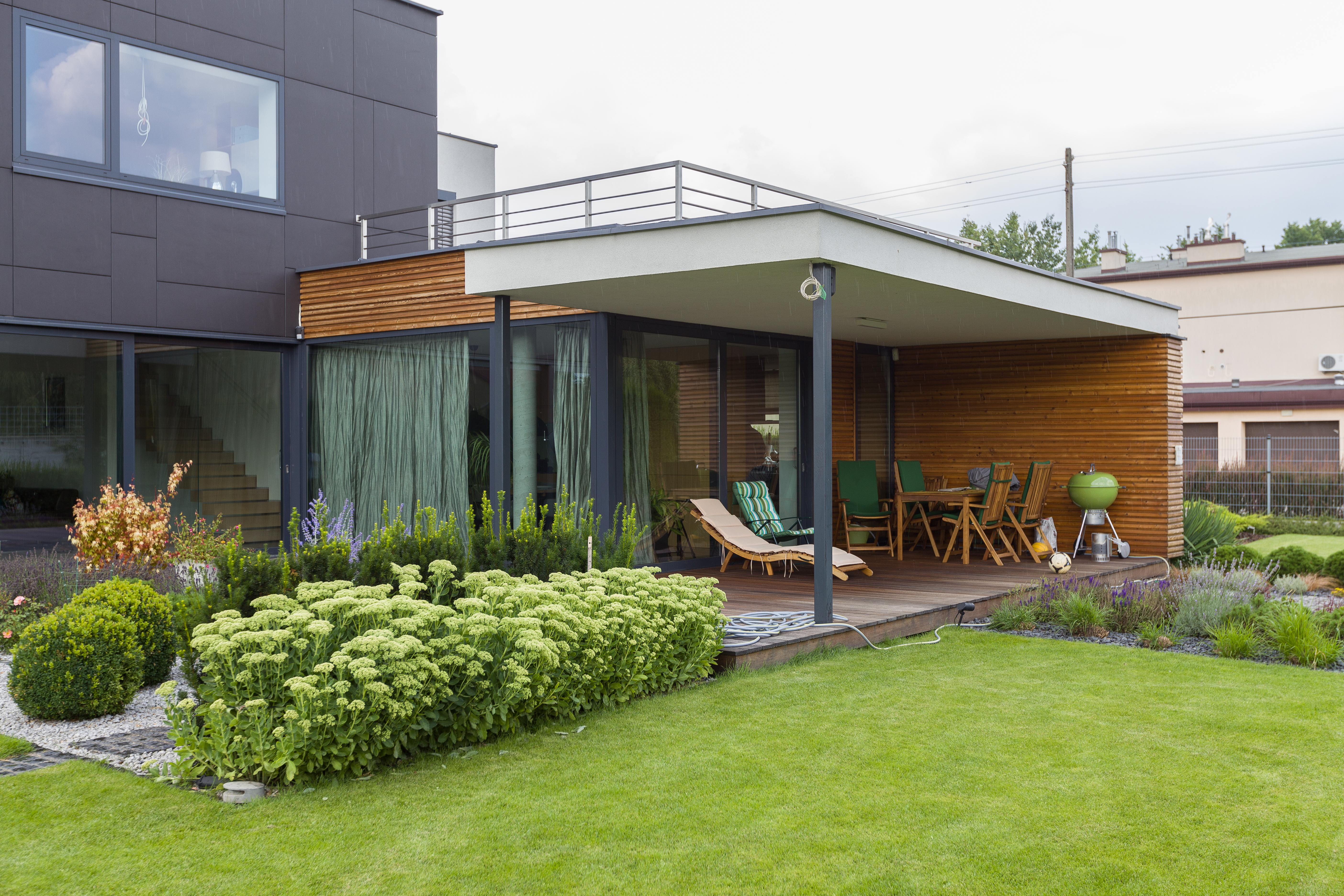 Wykończenie tarasu pod klucz z drewnianą podłogą i ścianami, meble ogrodowe na zadaszonym tarasie nowoczesnego domu o ciemnej elewacji.