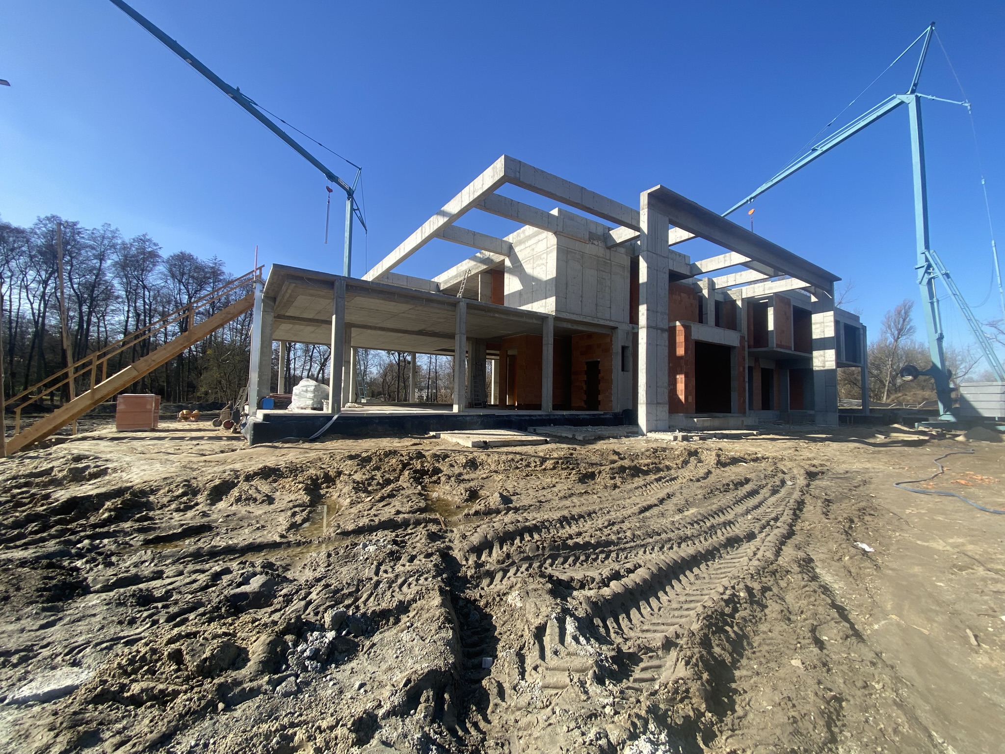 Zaawansowany etap budowy domu w stanie surowym z widoczną konstrukcją żelbetową i dźwigami na placu budowy.