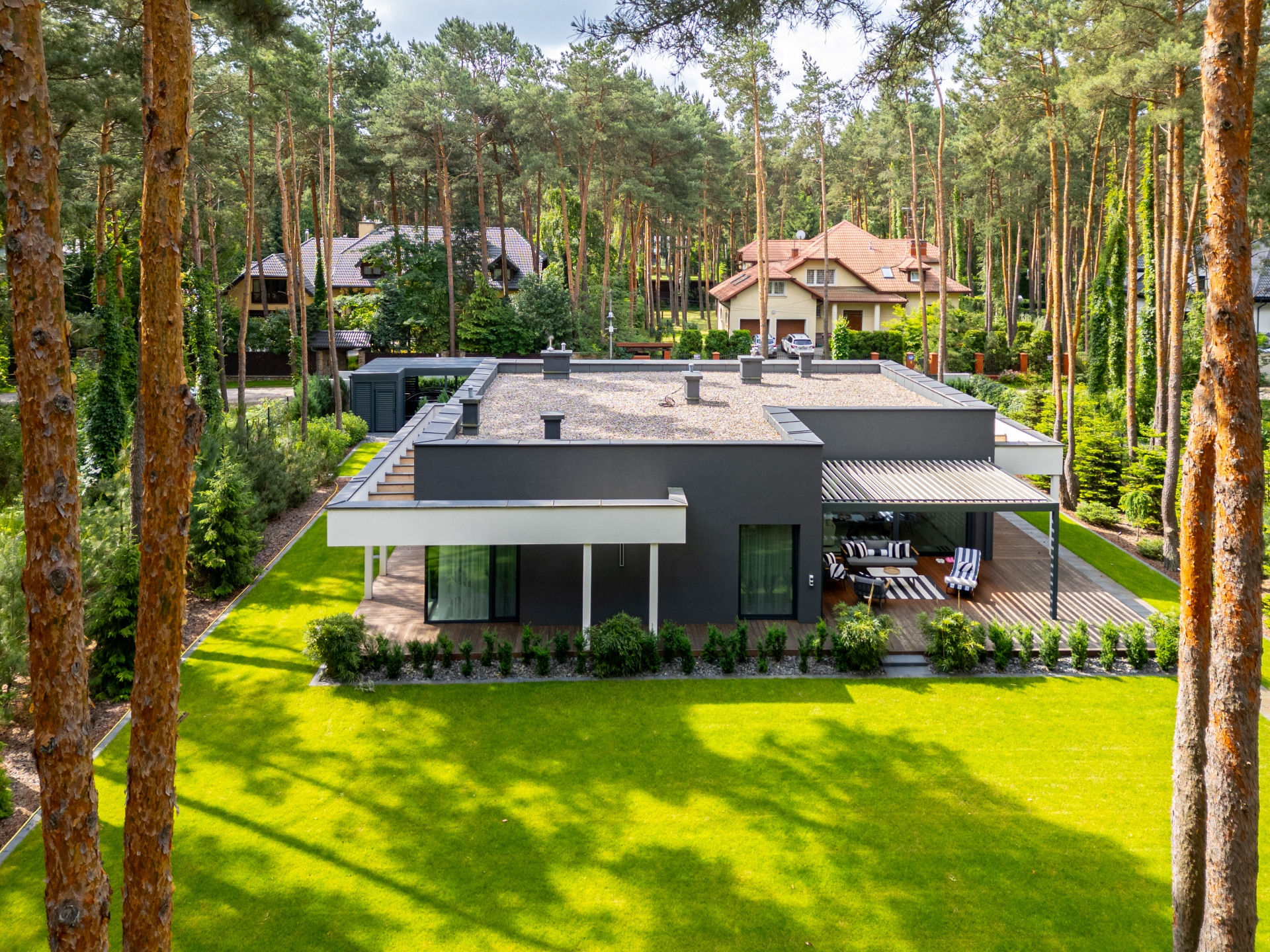 Luksusowy dom o grafitowej elewacji wkomponowany w starodrzew, zrealizowany przez New-House. Widok z góry na płaski dach z attyką, drewniany taras wypoczynkowy i perfekcyjnie utrzymaną zieleń.