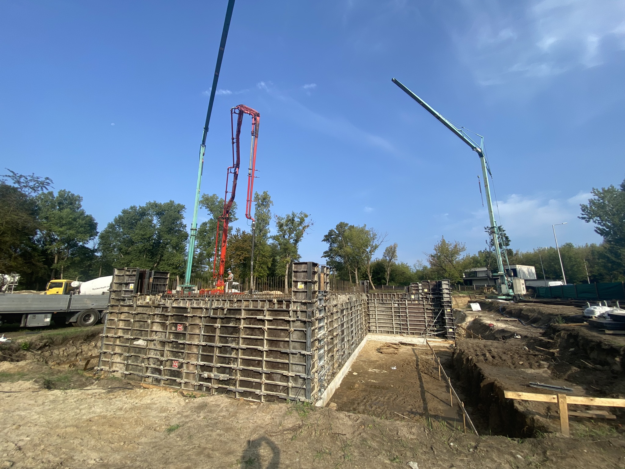 Plac budowy domu jednorodzinnego w słoneczny dzień. Na pierwszym planie widać metalowe deskowania systemowe przygotowane do betonowania ścian fundamentowych lub piwnicy. W tle pracują dwie pompy do betonu oraz dźwig na tle drzew i błękitnego nieba. Budowa prowadzona przez firmą budowlaną New-House