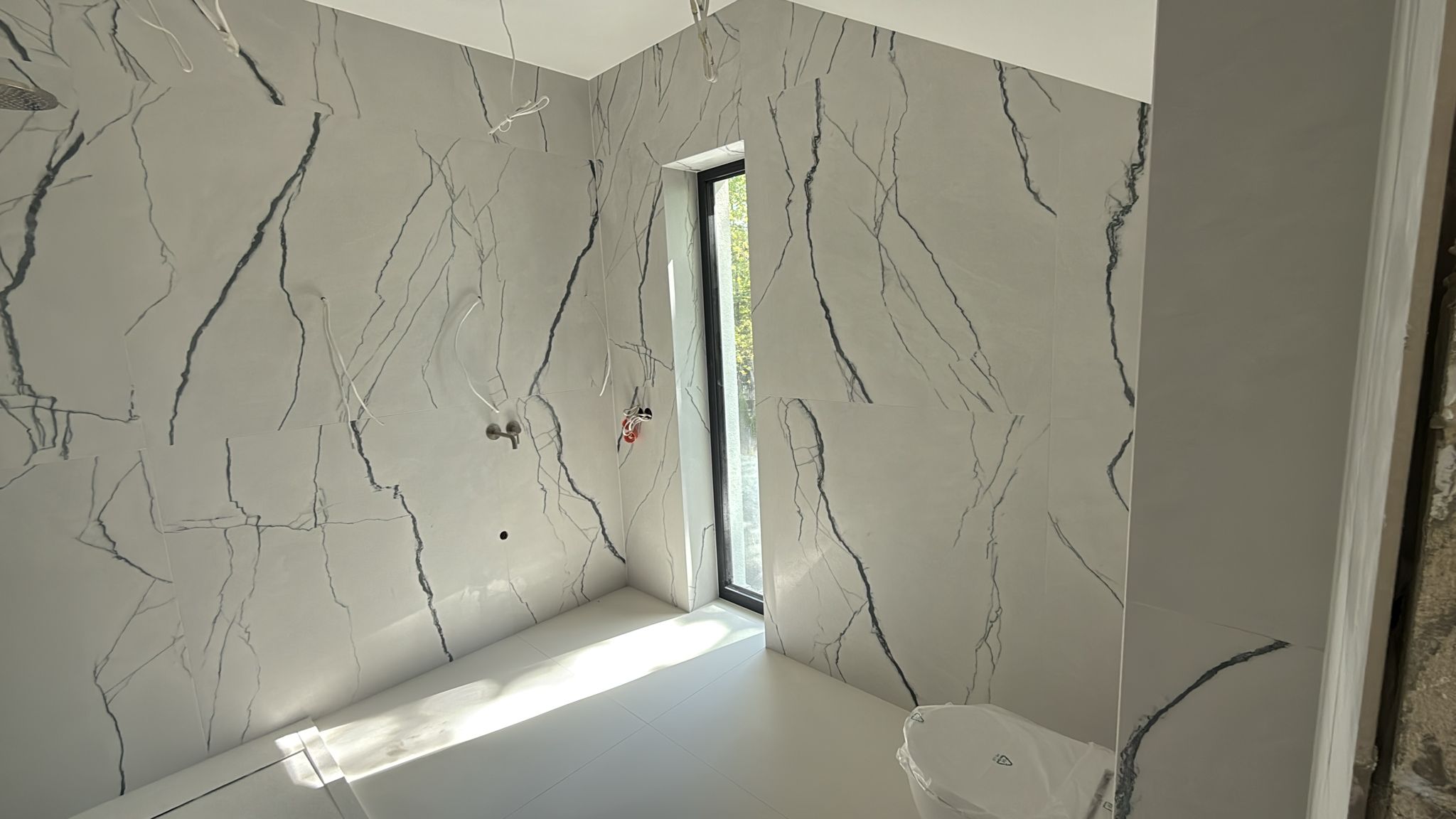 Wnętrze nowoczesnej łazienki w trakcie prac wykończeniowych. Ściany pokryte są wielkoformatowymi spiekami kwarcowymi w kolorze białym z wyraźnym, ciemnym żyłkowaniem imitującym marmur. Widoczne są wyprowadzenia instalacji pod baterie podtynkowe oraz wąskie, wysokie okno wpuszczające naturalne światło. Realizacja New-House