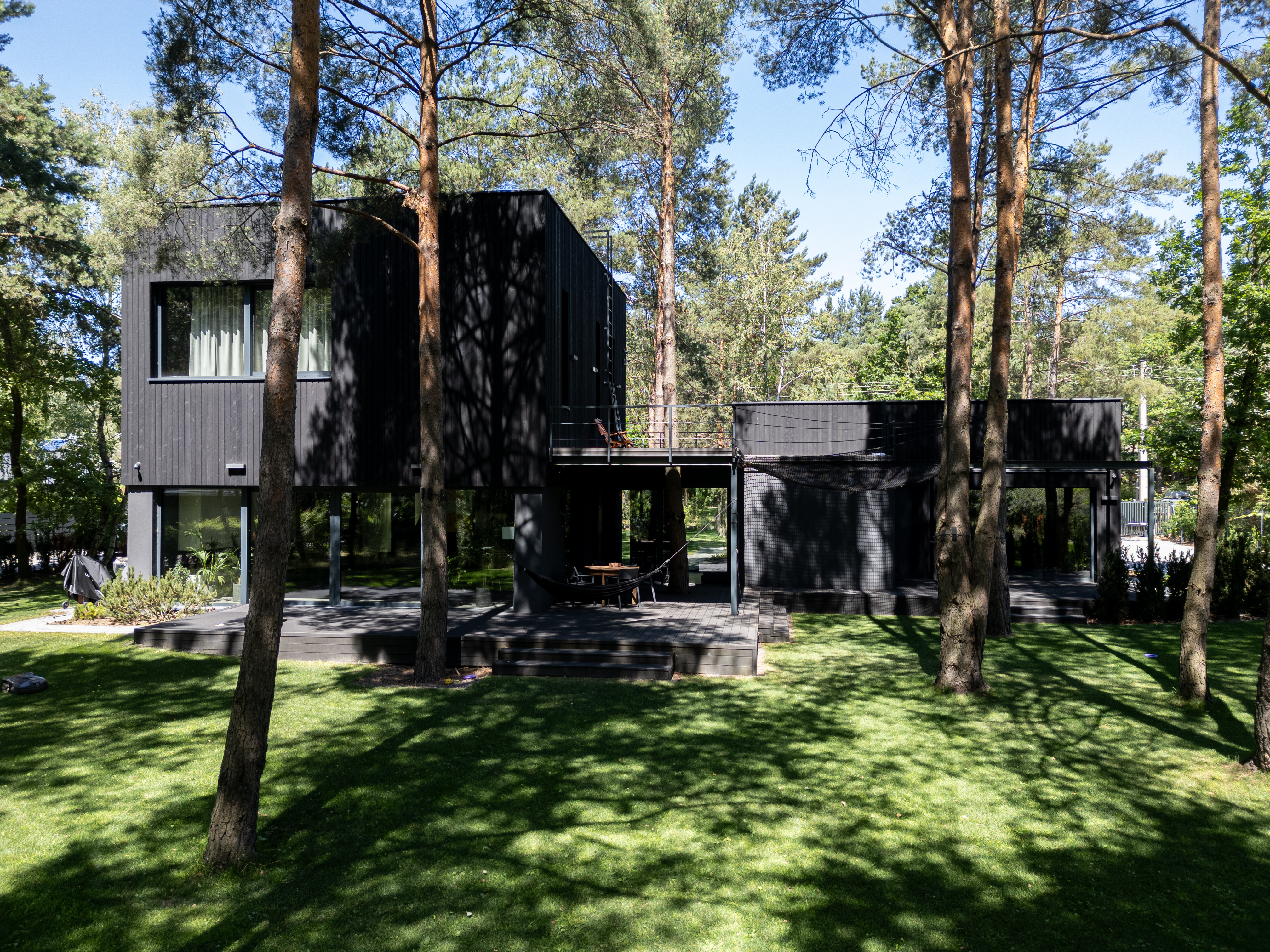 Nowoczesny dom piętrowy o czarnej, drewnianej elewacji, wkomponowany w gęsty las sosnowy. Budynek posiada duże przeszklenia na parterze oraz przestronny, ciemny taras z meblami wypoczynkowymi i hamakiem. Konstrukcja charakteryzuje się minimalistyczną, geometryczną bryłą. Wykonanie New-House